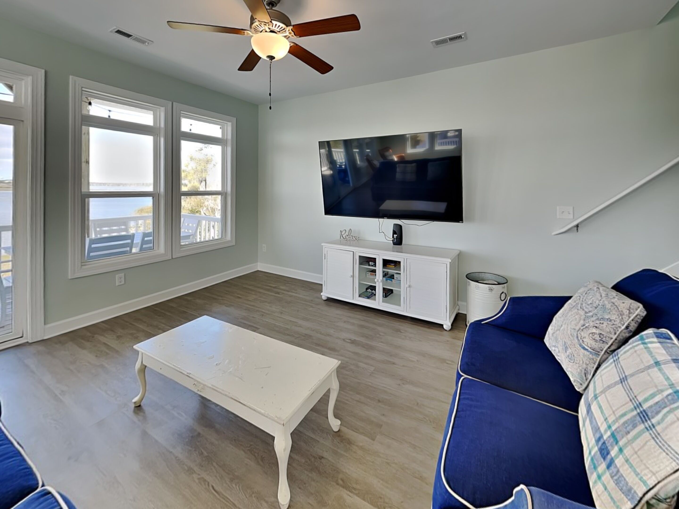 Una sala de estar con ventilador de techo, televisor, mueble blanco, sofá azul y ventana con vista.