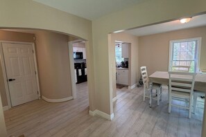 Een ruime woonkamer met een eettafel en stoelen, een keuken op de achtergrond en een deur naar een andere kamer.