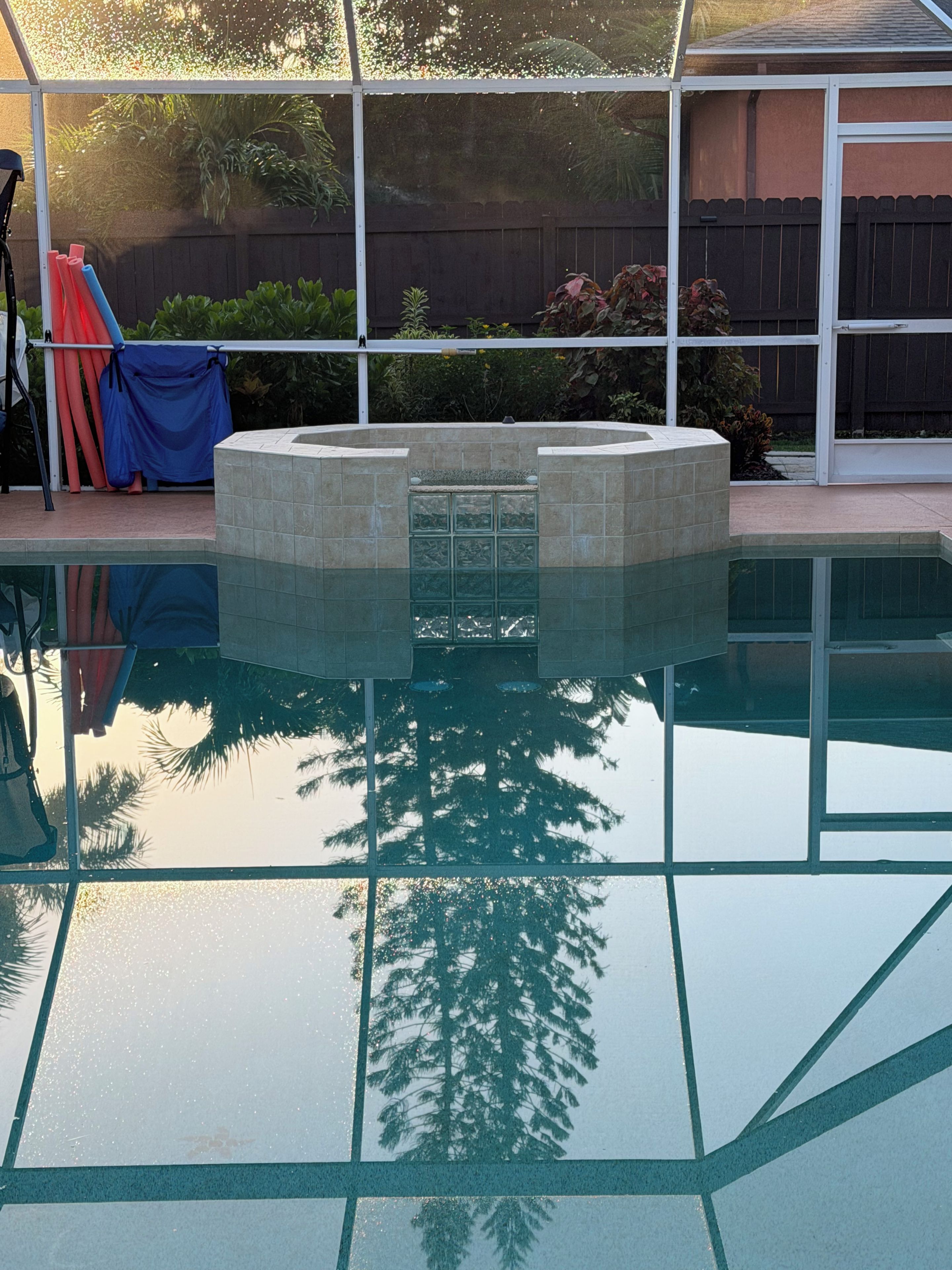 A swimming pool with a tiled deck, surrounded by a fence and vegetation.