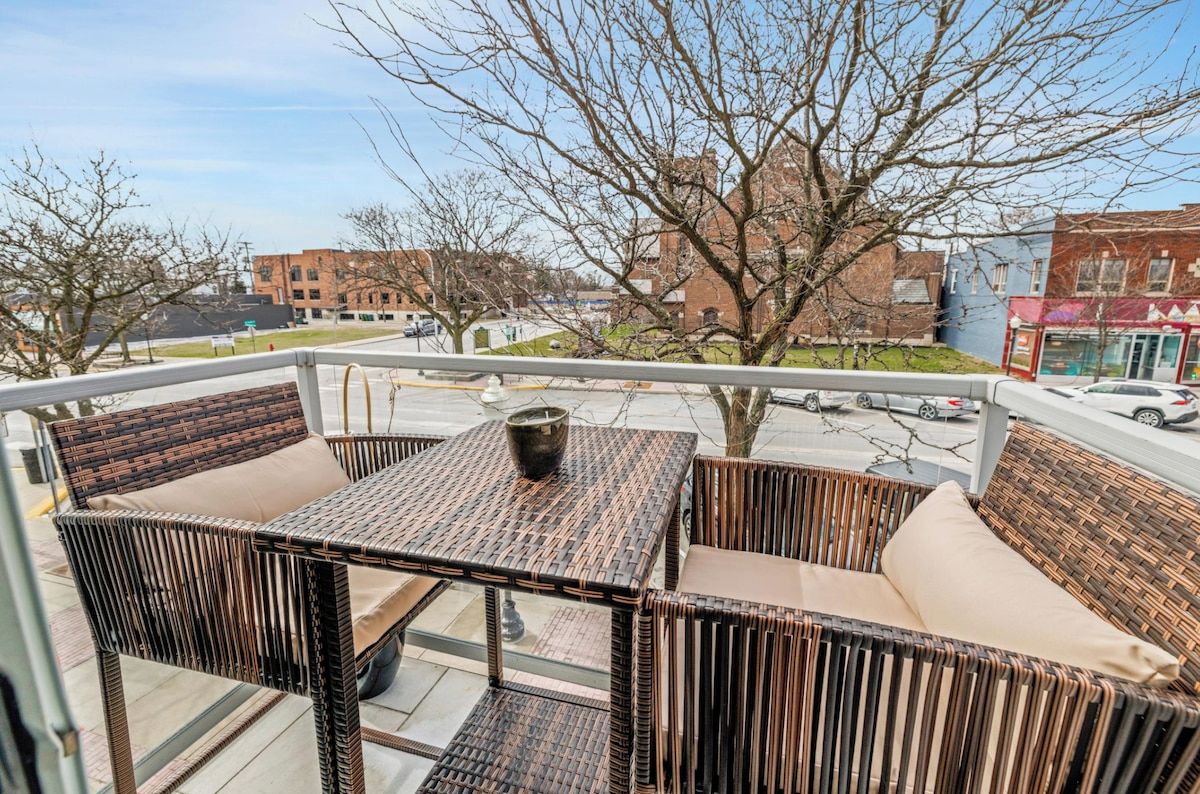 A balcony with wicker furniture, a glass table, and a view of the street and buildings.