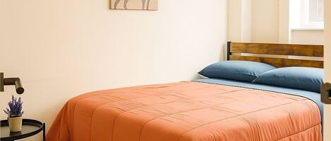 A bedroom with a bed, a small table with a plant, a wall-mounted painting, and a window.