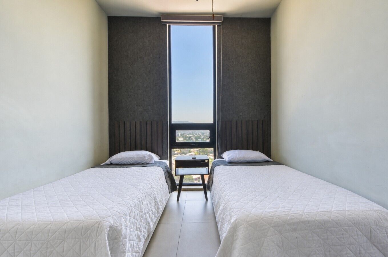 A modern bedroom with two beds, a view of the city through a large window, and a small table between the beds.