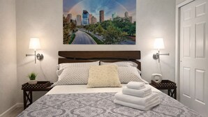 A hotel room with a bed, two bedside tables, a nightstand, a lamp, a painting of a cityscape, and a door.