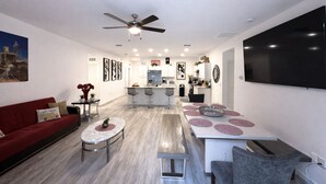 A modern living room with a red sofa, a round coffee table, a dining area with a large table and bar stools, a kitchen with white cabinets, and a ceiling fan.