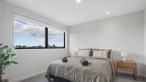 A bedroom with a large window, a bed with pillows, a bedside table with a lamp, and a plant. - Waihi Beach Retreat (#B7) (Waihi Beach)