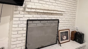 Una habitación con chimenea de ladrillo blanco, un televisor montado y un cuadro en el repisa.