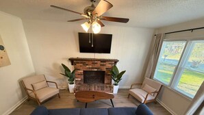A living room with a dark blue sofa, two beige armchairs, a wooden coffee table, a brick fireplace, a ceiling fan, a mounted TV, and a large window with blinds. - Newly Renovated 3 Bedroom House in Lakewood (Durham)