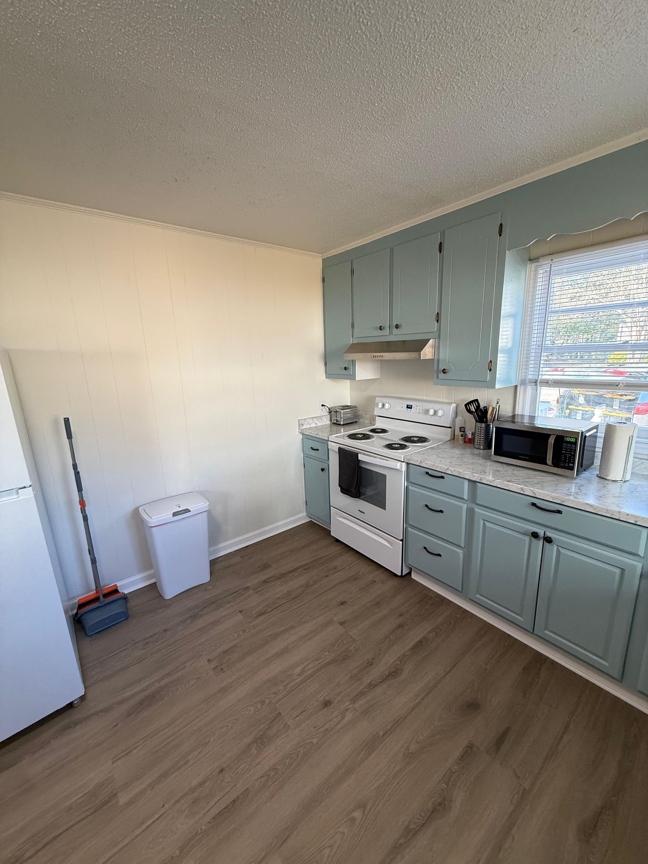 A kitchen with light blue cabinets, a white stove, a microwave, and a trash bin.