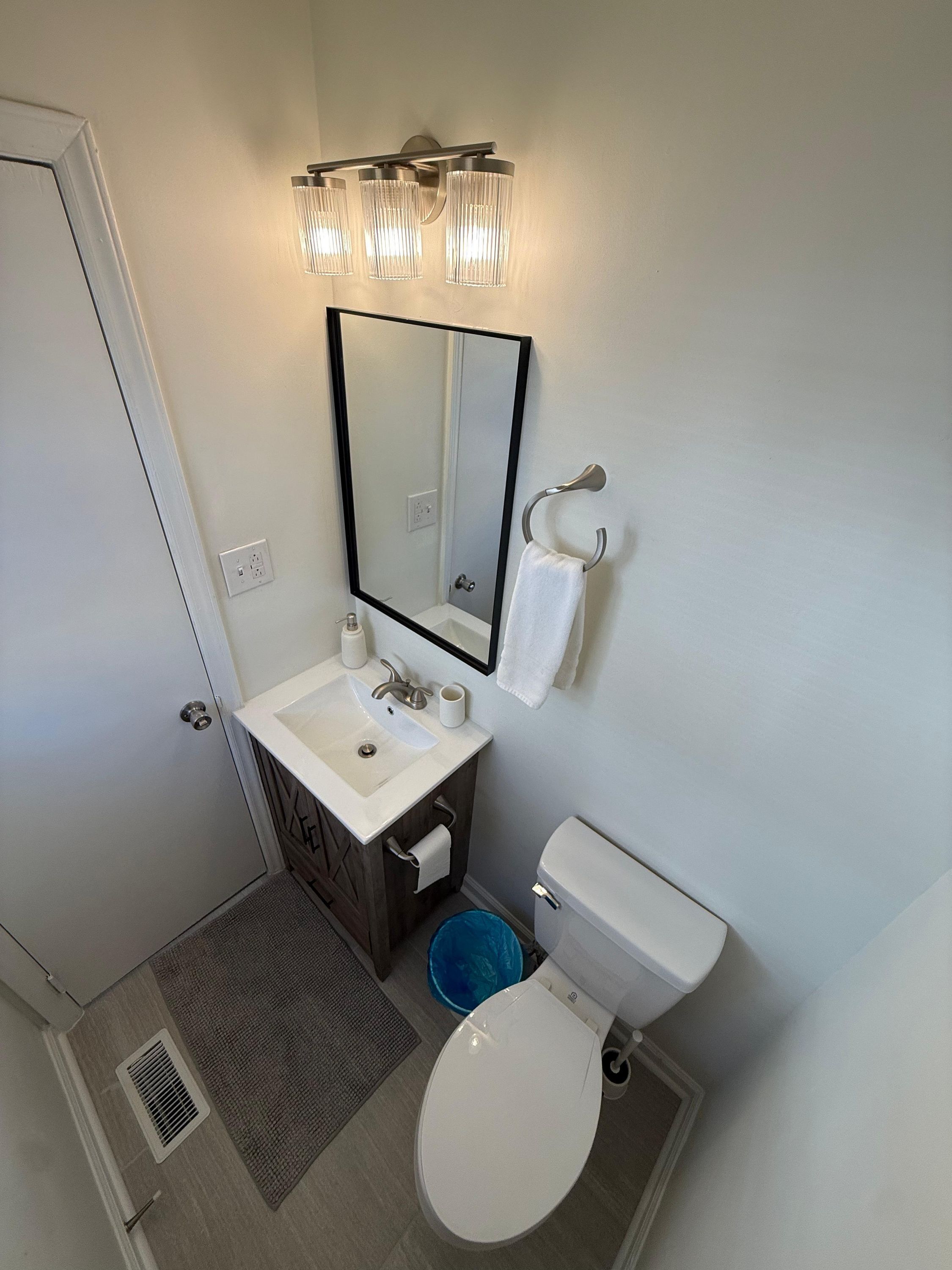 Ein Badezimmer mit einem weißen Waschbecken, einem Spiegel und einer Toilette. Ein Handtuchhalter und ein blauer Mülleimer sind ebenfalls vorhanden.