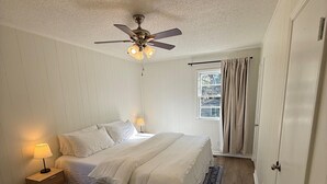 A bedroom with a bed, a ceiling fan, a nightstand with a lamp, a window with curtains, and a door. - Newly Renovated 3 Bedroom House in Lakewood (Durham)