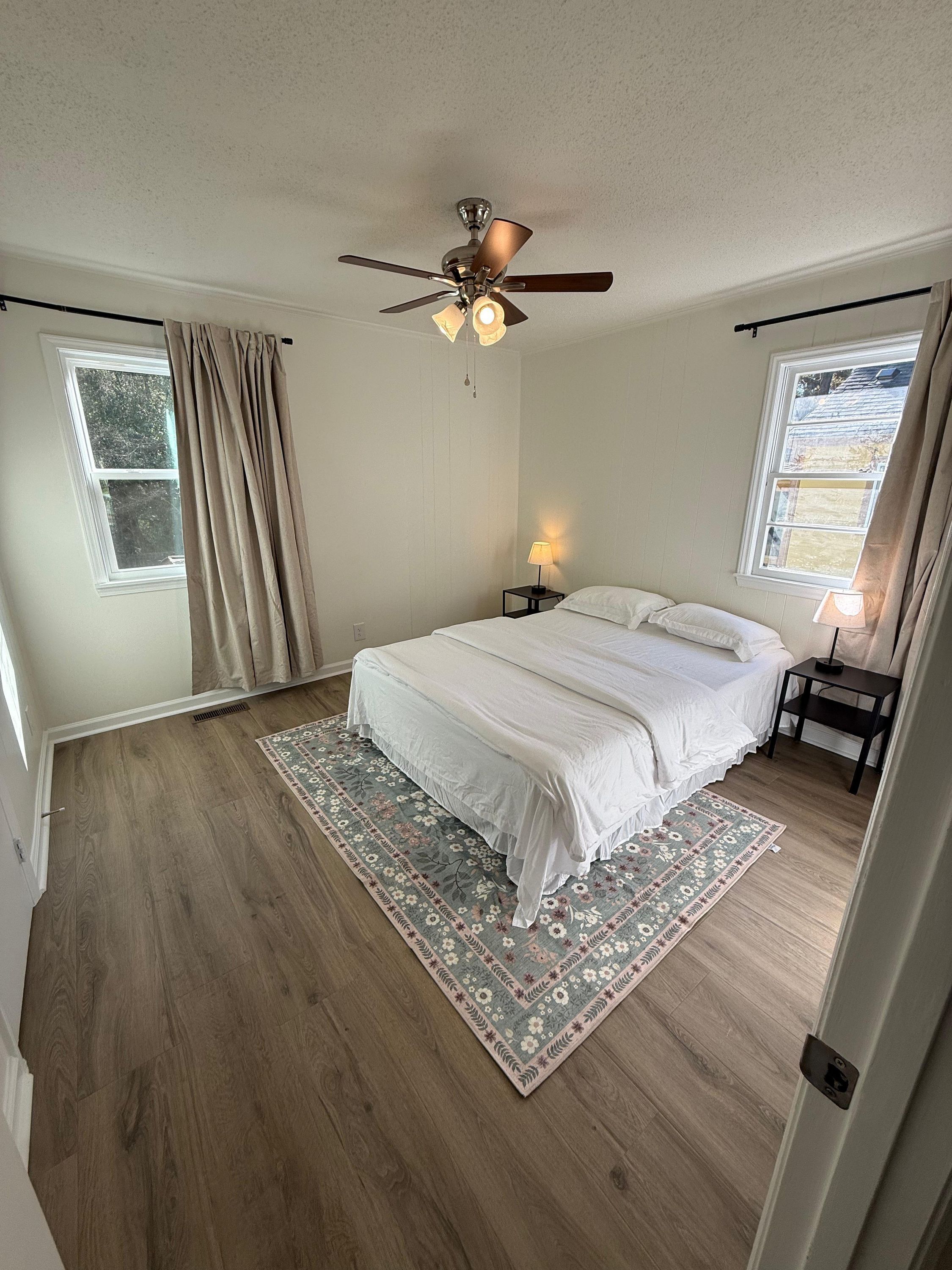 A bedroom with a bed, a ceiling fan, two bedside tables with lamps, a window with curtains, and a wooden floor.