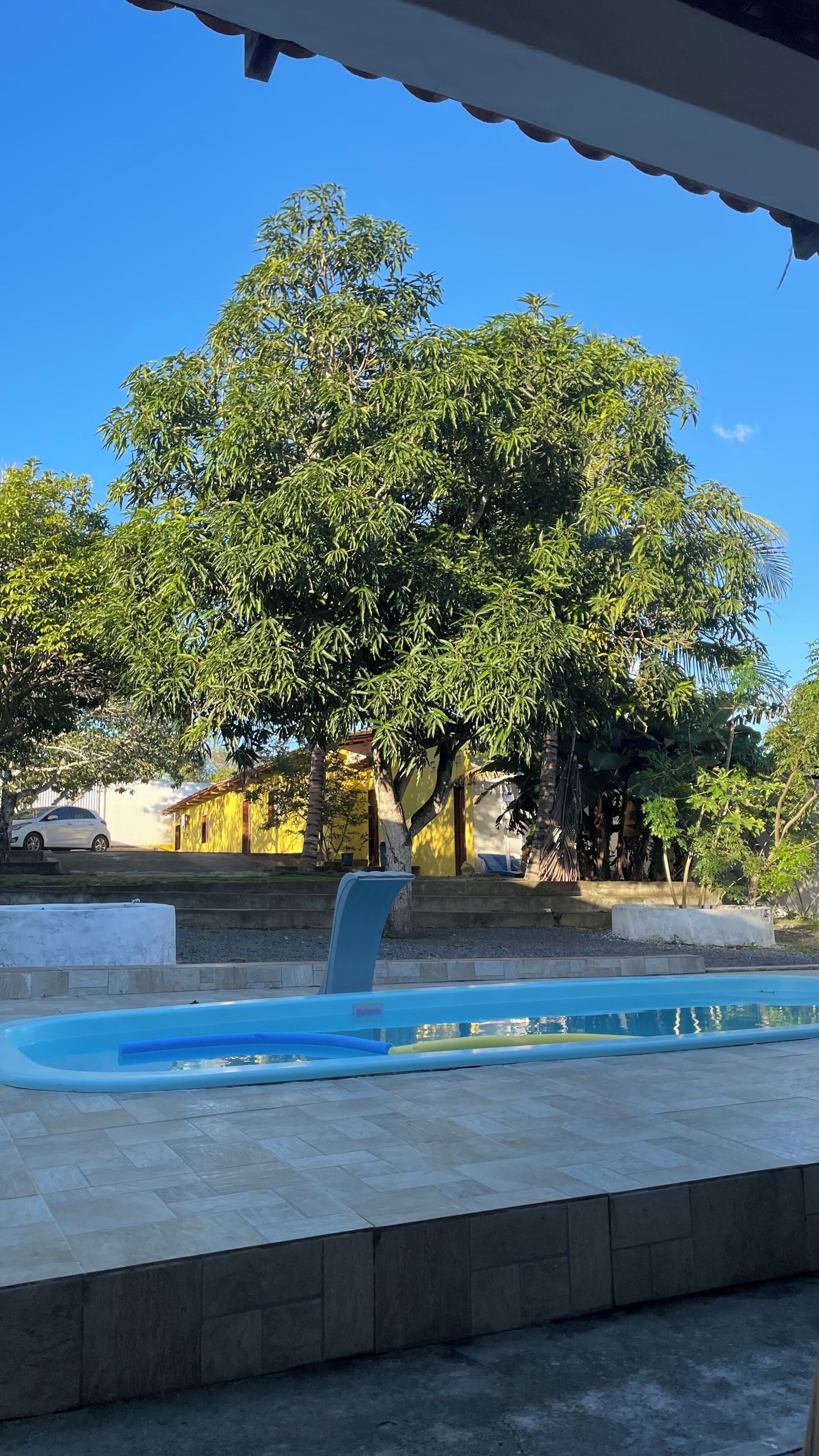 Piscina com céu azul límpido ao fundo, rodeada por vegetação e um edifício com fachada amarela.