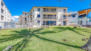 Un immeuble résidentiel moderne à plusieurs étages, doté de balcons, d’une pelouse bien entretenue et surplombant un ciel bleu dégagé.