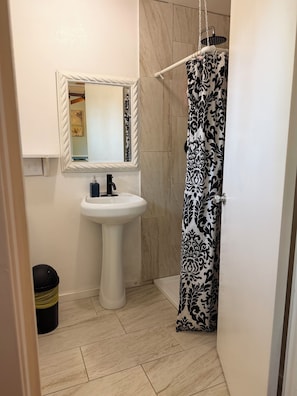 A bathroom with a pedestal sink, a mirror, a shower curtain, and a trash bin.