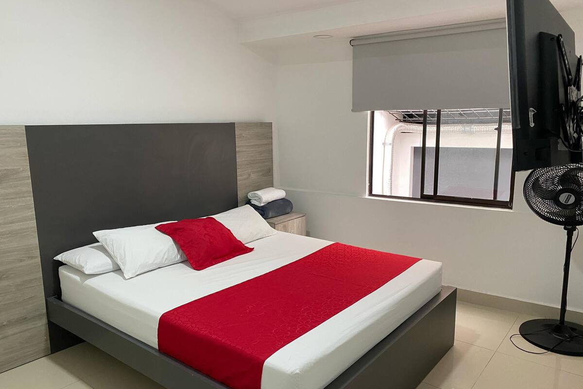 Une chambre moderne avec un lit, une table de chevet, une fenêtre avec des stores et un ventilateur sur pied.