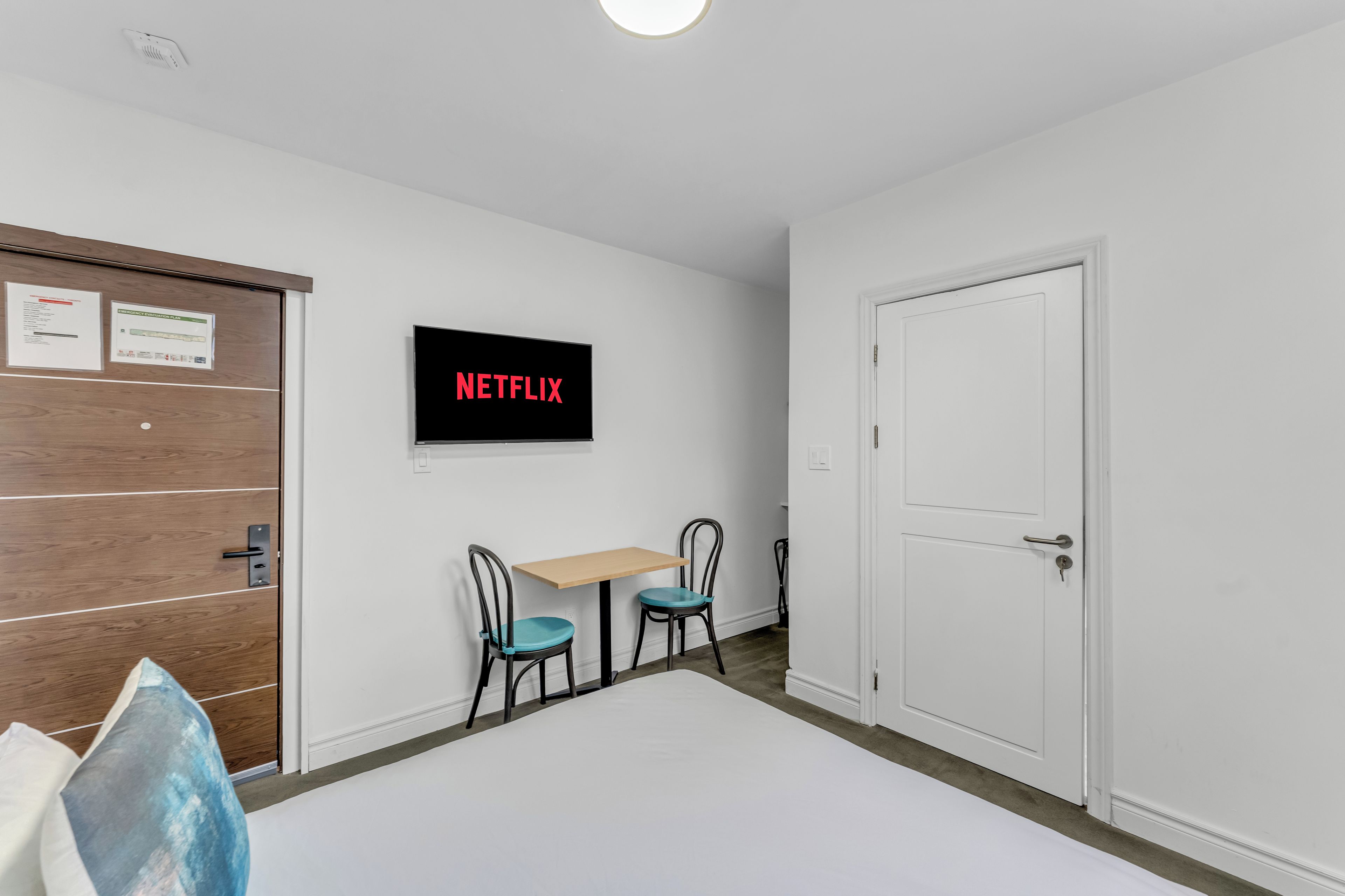 Una habitación con una puerta blanca, una puerta de madera, una mesa con dos sillas y un televisor montado en la pared que muestra 'NETFLIX'.
