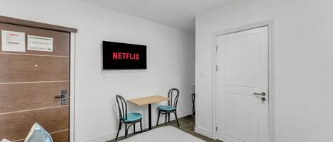Una habitación con una puerta blanca, una puerta de madera, una mesa con dos sillas y un televisor montado en la pared que muestra 'NETFLIX'.