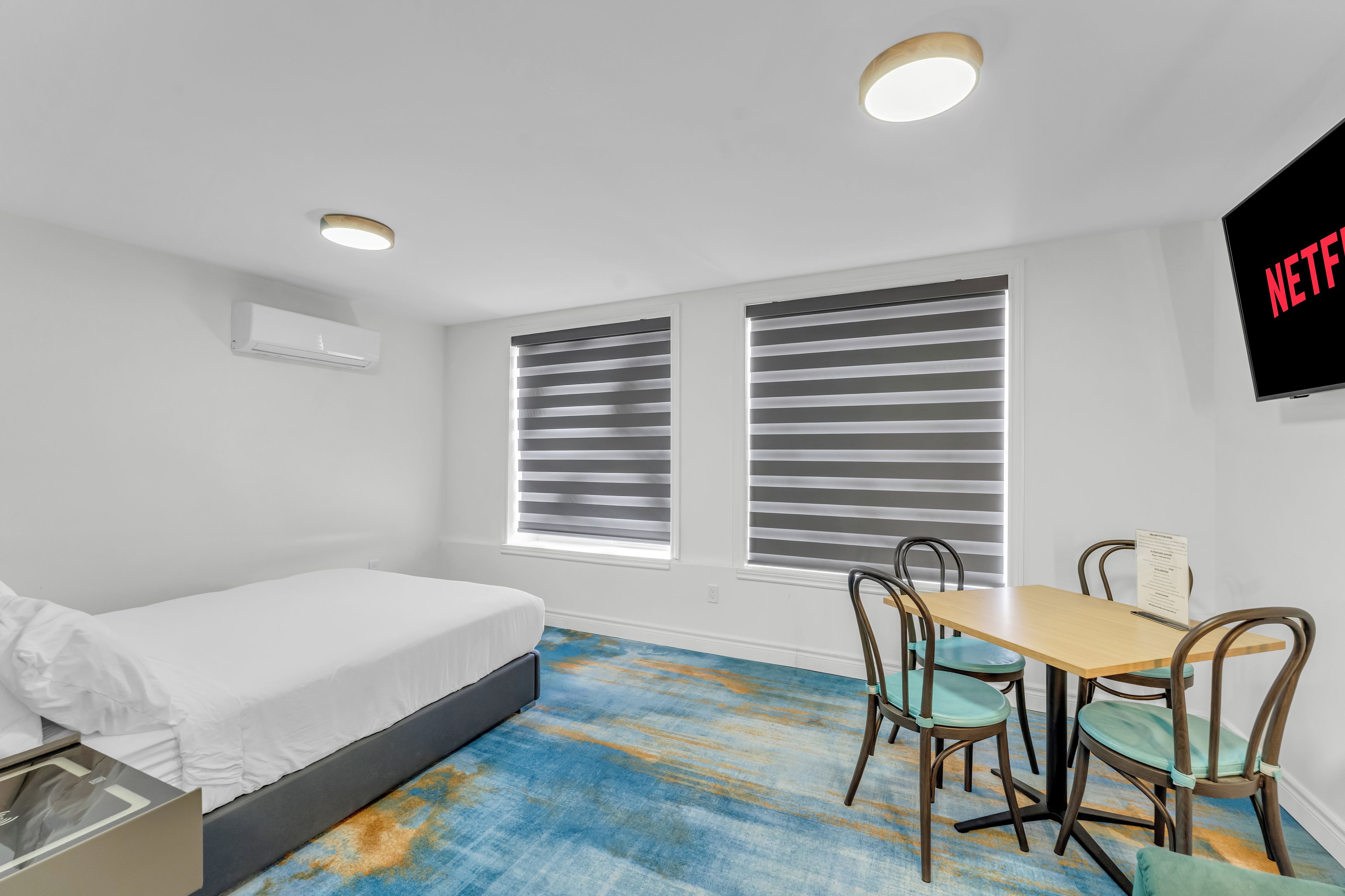 A room with a bed, a table with chairs, a TV mounted on the wall, and a window with blinds.