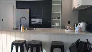 A modern kitchen with a white countertop, black cabinets, and a built-in oven. There are three black stools and a coffee maker on the counter. - chevron /3beds / Pool /Parking n Ocean glimpses (Surfers Paradise)