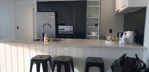 A modern kitchen with a white countertop, black cabinets, and a built-in oven. There are three black stools and a coffee maker on the counter. - chevron /3beds / Pool /Parking n Ocean glimpses (Surfers Paradise)