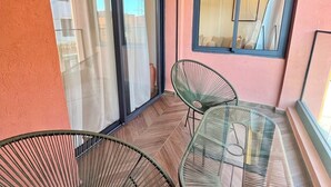 A balcony with two green wire-framed chairs and a glass table, a sliding glass door, and a window with a view of a building. - Cocon Plaza (Marrakech)