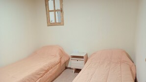 Una habitación pequeña y sencilla con dos camas individuales, una mesita de noche de madera y una ventana.