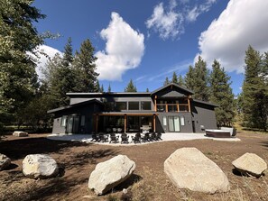 A modern two-story lodge with a covered patio area, surrounded by large rocks and dense forest. - Ridge House- new home, sleeps 16, quiet neighborhood-, hot tub, trailer parking, pet friendly (McCall)