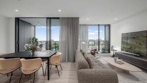 A modern living room with a large TV, a sofa, a dining table with chairs, and a view of the city through large windows. - Ocean Glimpse - A 3-Bedroom Designer Sub-Penthouse @ 333STK (Port Phillip)