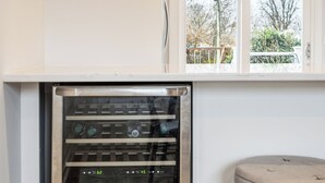Et lille, stålfarvet minikøleskab med flere hylder, placeret i et hjørne med trægulv og et rundt bord i nærheden.