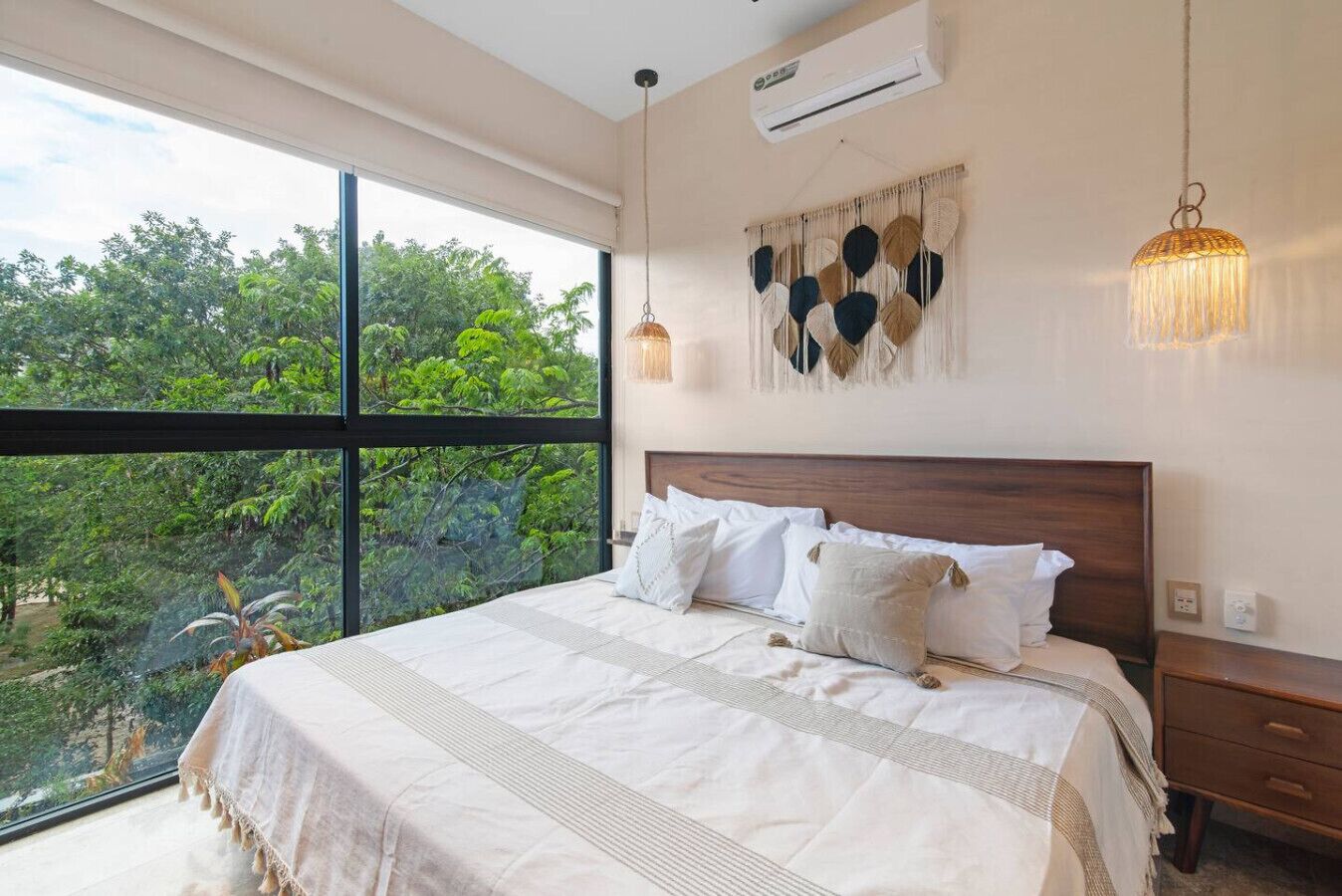 A bedroom with a large window, a bed with white bedding, a wooden headboard, and a wall-mounted air conditioner.