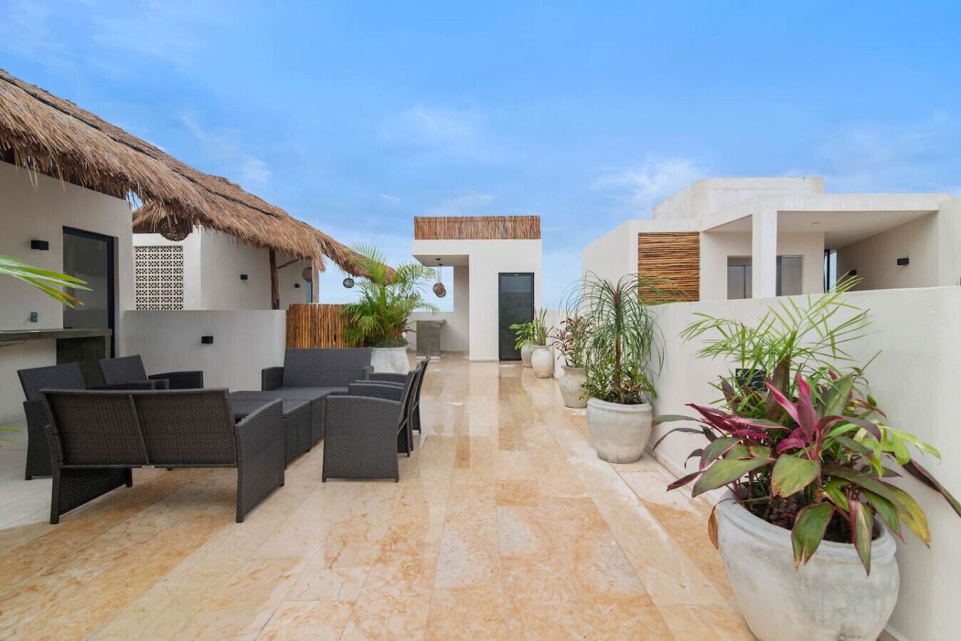 A sunlit outdoor patio with wicker furniture, potted plants, and a thatched roof structure.