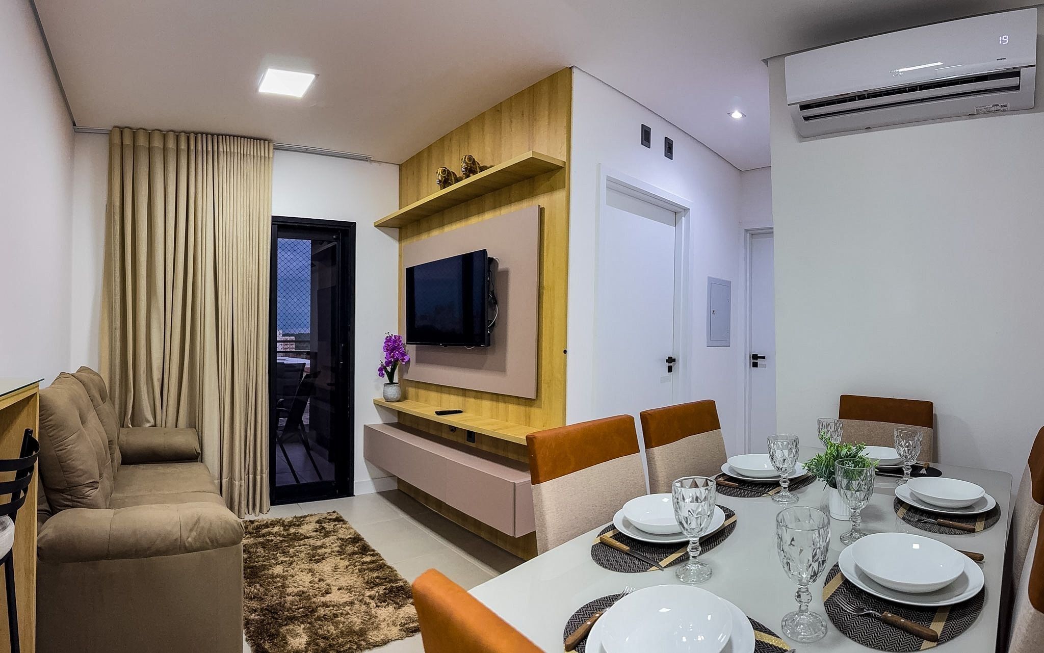 A modern living room with a sofa, dining table set for four, a flat-screen TV mounted on the wall, and an air conditioning unit.