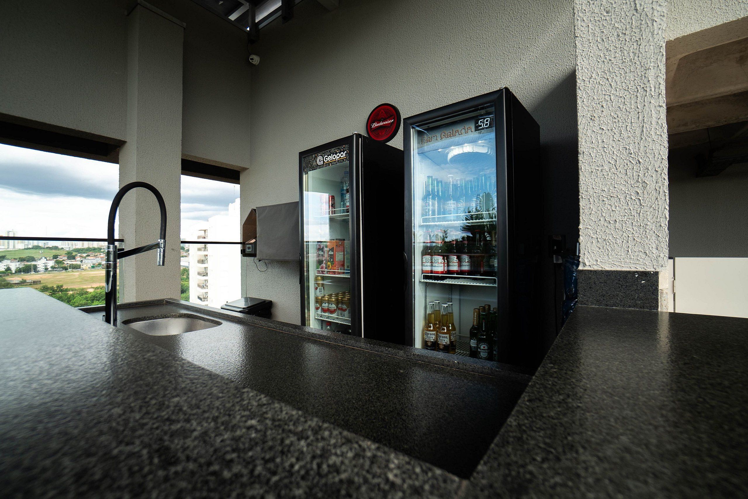 Una cocina con dos refrigeradores llenos de bebidas, un lavamanos y un ventanal con vistas a un paisaje urbano.