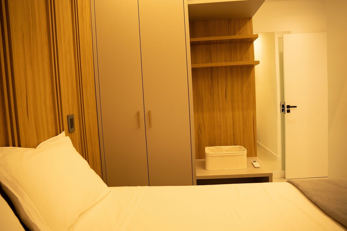 A bedroom with a bed, a wardrobe, a shelf unit, and a bedside table with a tissue box and a remote control.