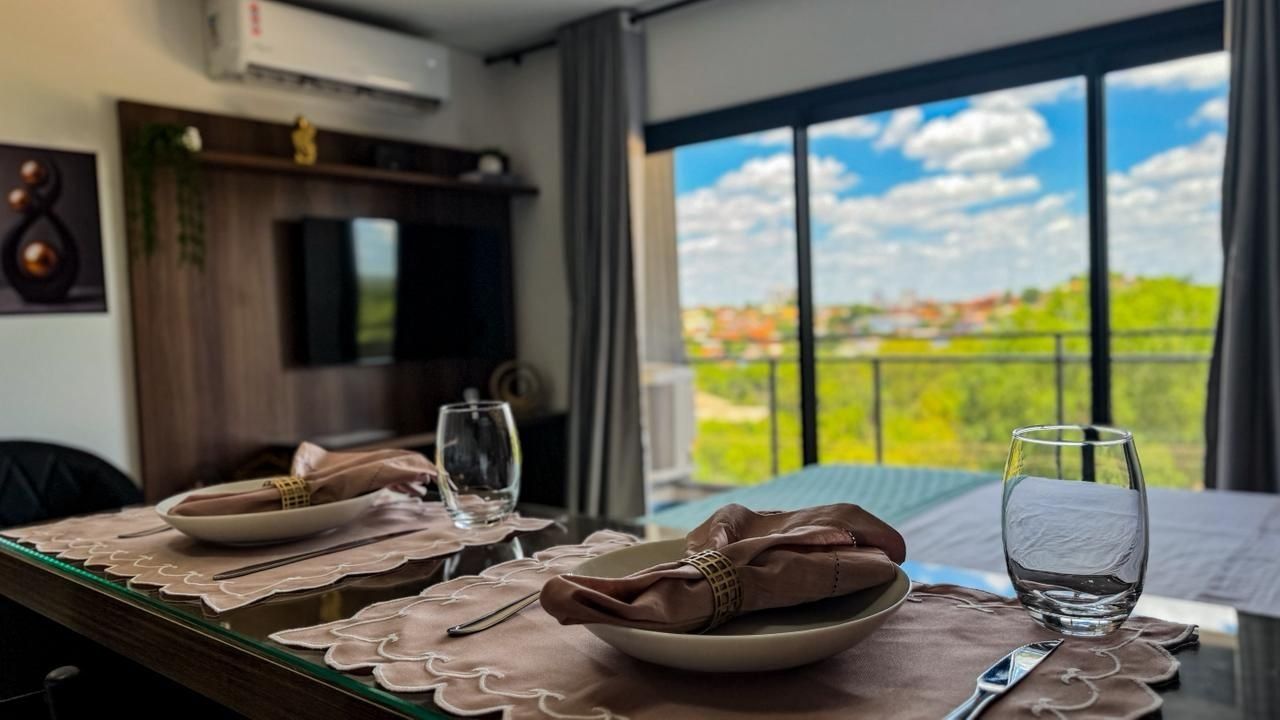 Mesa lista con platos, vasos y servilleta, con vista a follaje y un edificio a través de una ventana.