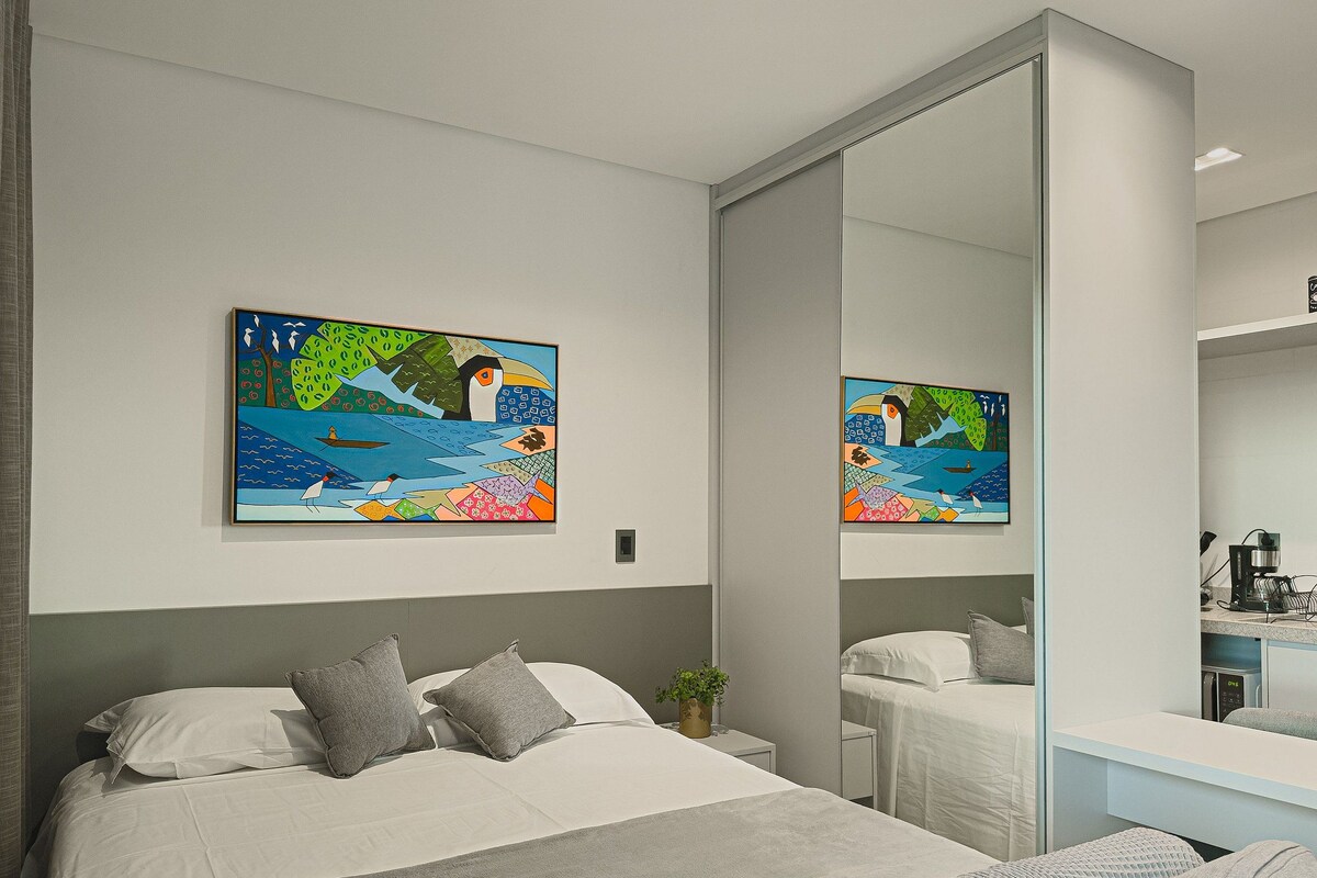 A bedroom with a bed, pillows, a bedside table with a plant, and a large mirror reflecting the room.