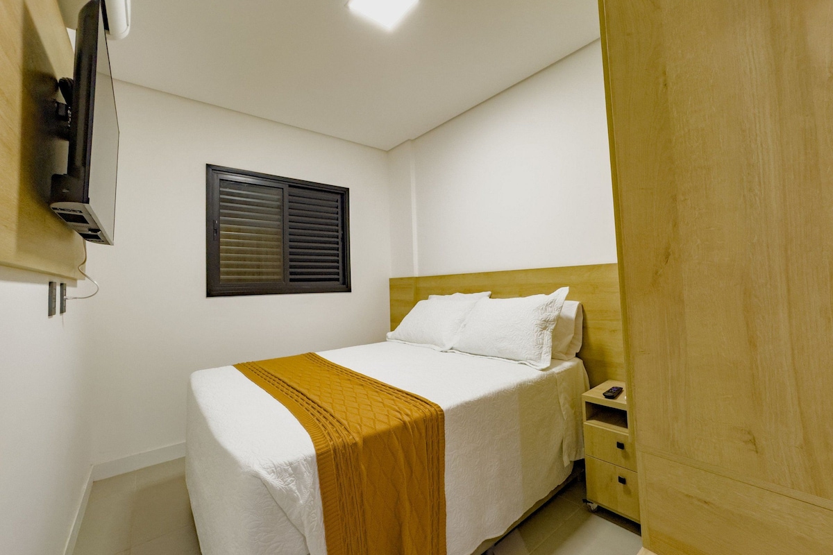 Un dormitorio con cama, mesita de noche, televisor montado en la pared y ventana con persianas.