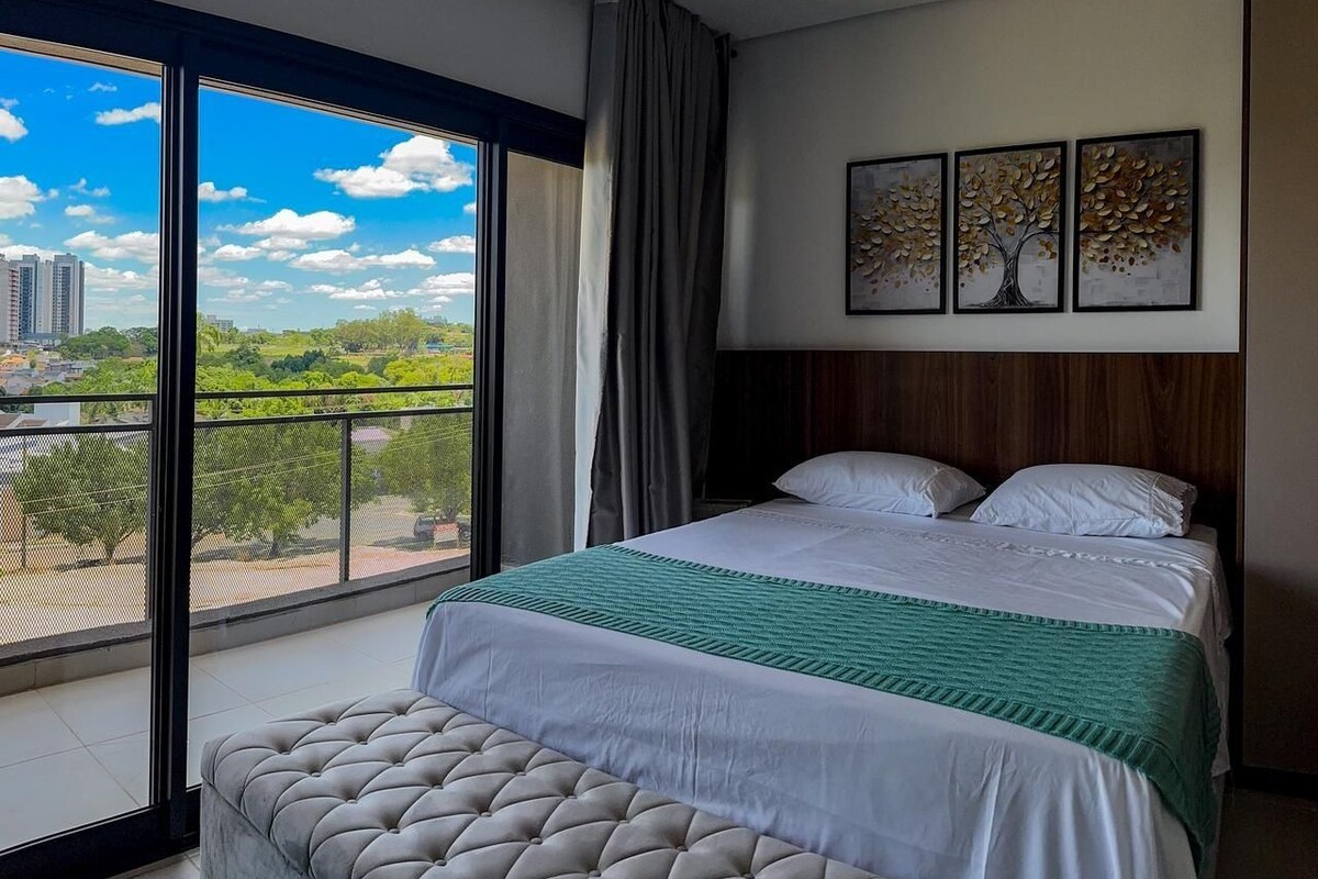 A hotel room with a large window, a bed with a green blanket, and a view of the cityscape.