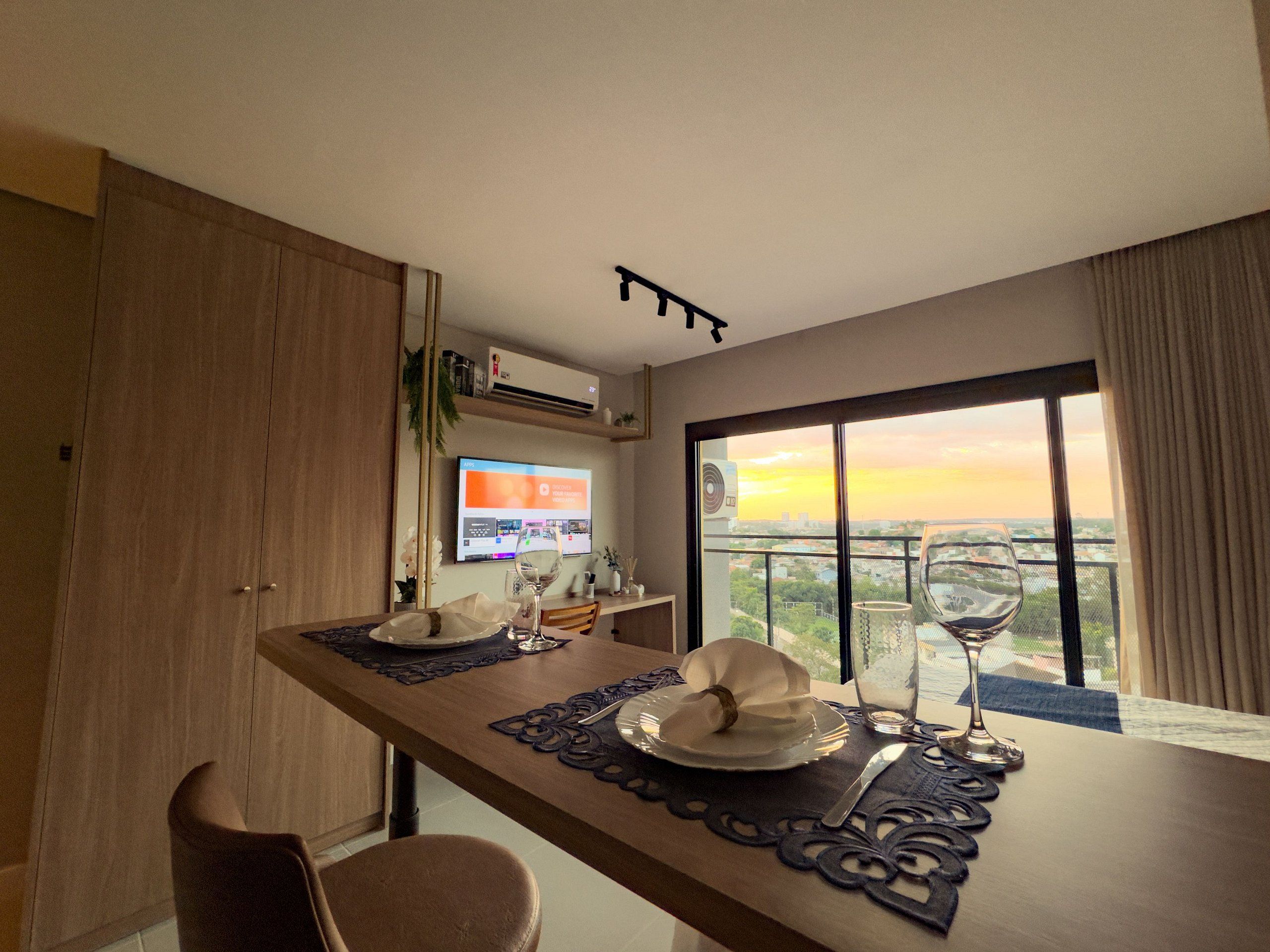 A dining area with a table set for two, a television mounted on the wall, and a large window with a view of the cityscape.