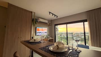 Et spiseområde med et bord dækket til to personer, et fjernsyn ophængt på væggen og et stort vindue med udsigt over bybilledet.