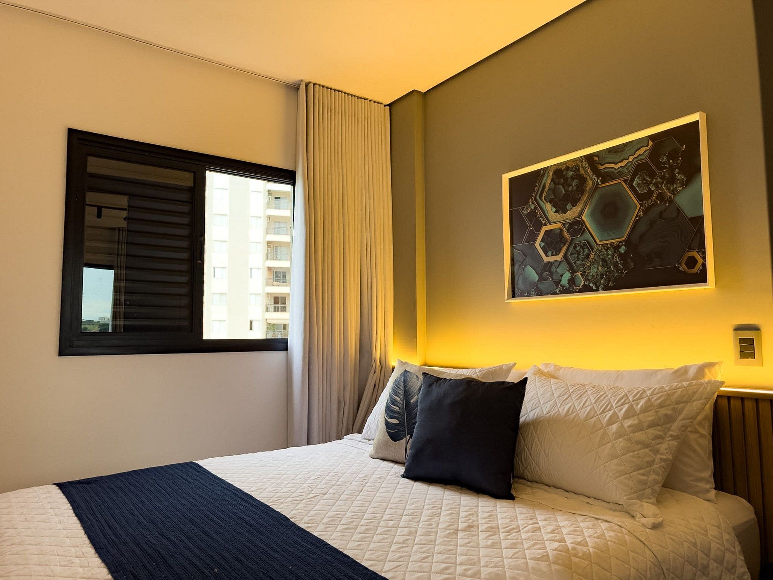 Una habitación de hotel con una cama, una ventana con persianas, una cortina, una obra de arte colgada en la pared y una mesita de noche.