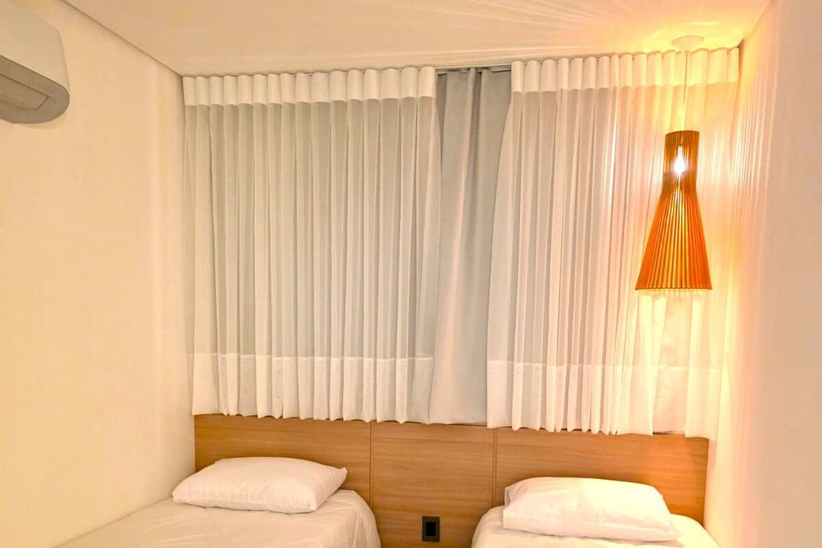 A hotel room with two single beds, a wooden headboard, white curtains, and a wall-mounted lamp.