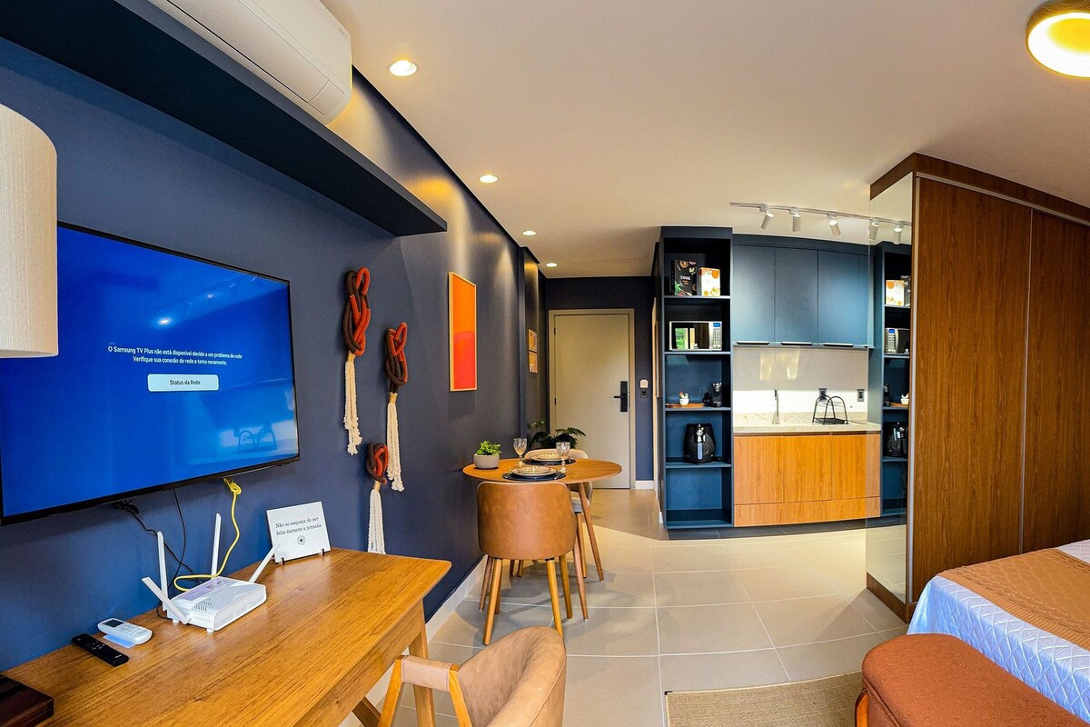 A modern hotel room with a flat-screen TV, a desk with a router, a bed with a wooden headboard, and a kitchenette.