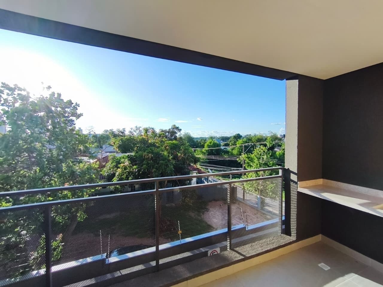A balcony with a metal railing, a white shelf, and a view of greenery and buildings.