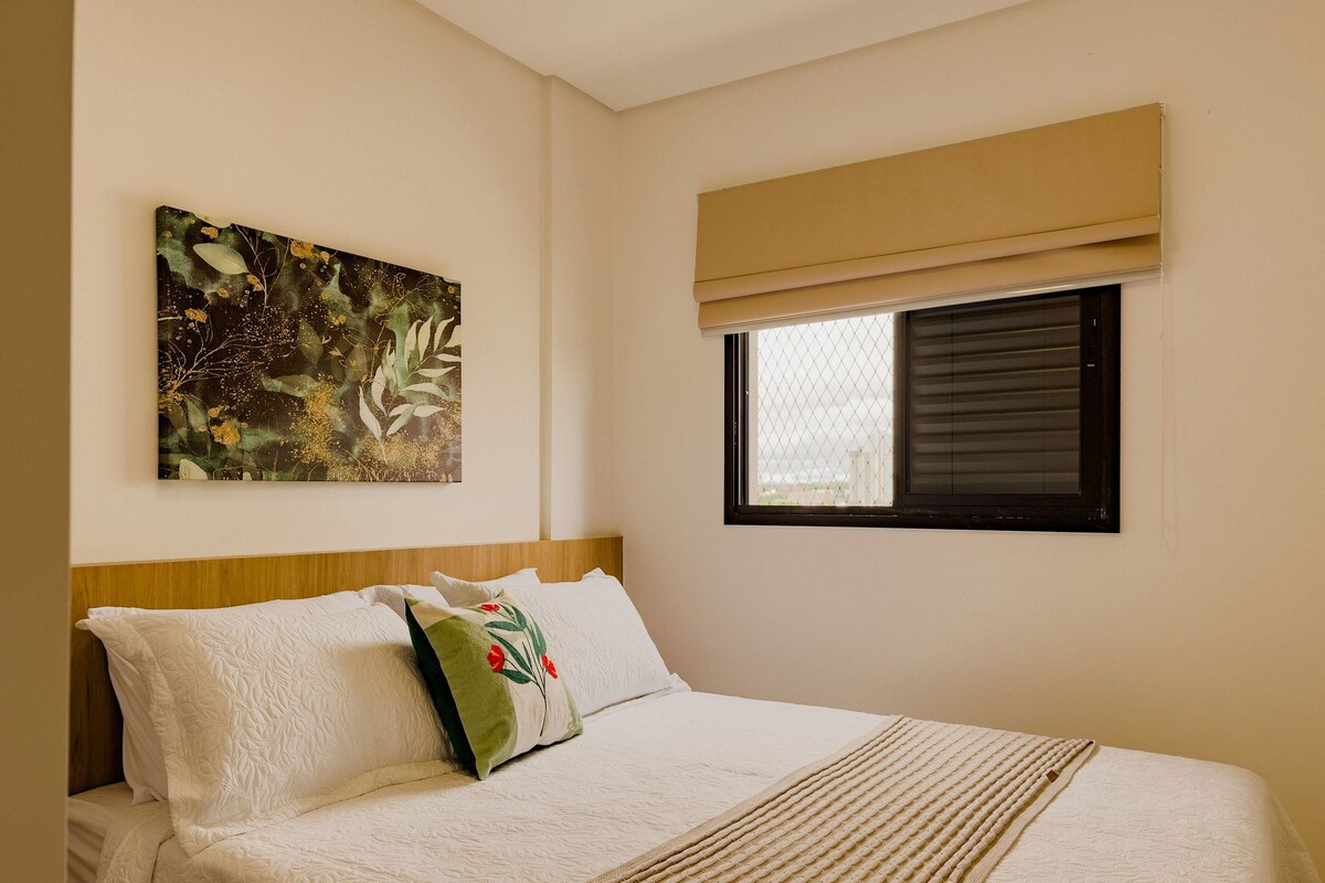 A bedroom with a bed, a window with blinds, a painting on the wall, and a headboard.