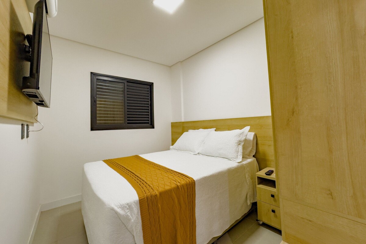 A bedroom with a bed, a bedside table, a nightstand, a television mounted on the wall, and a window with blinds.