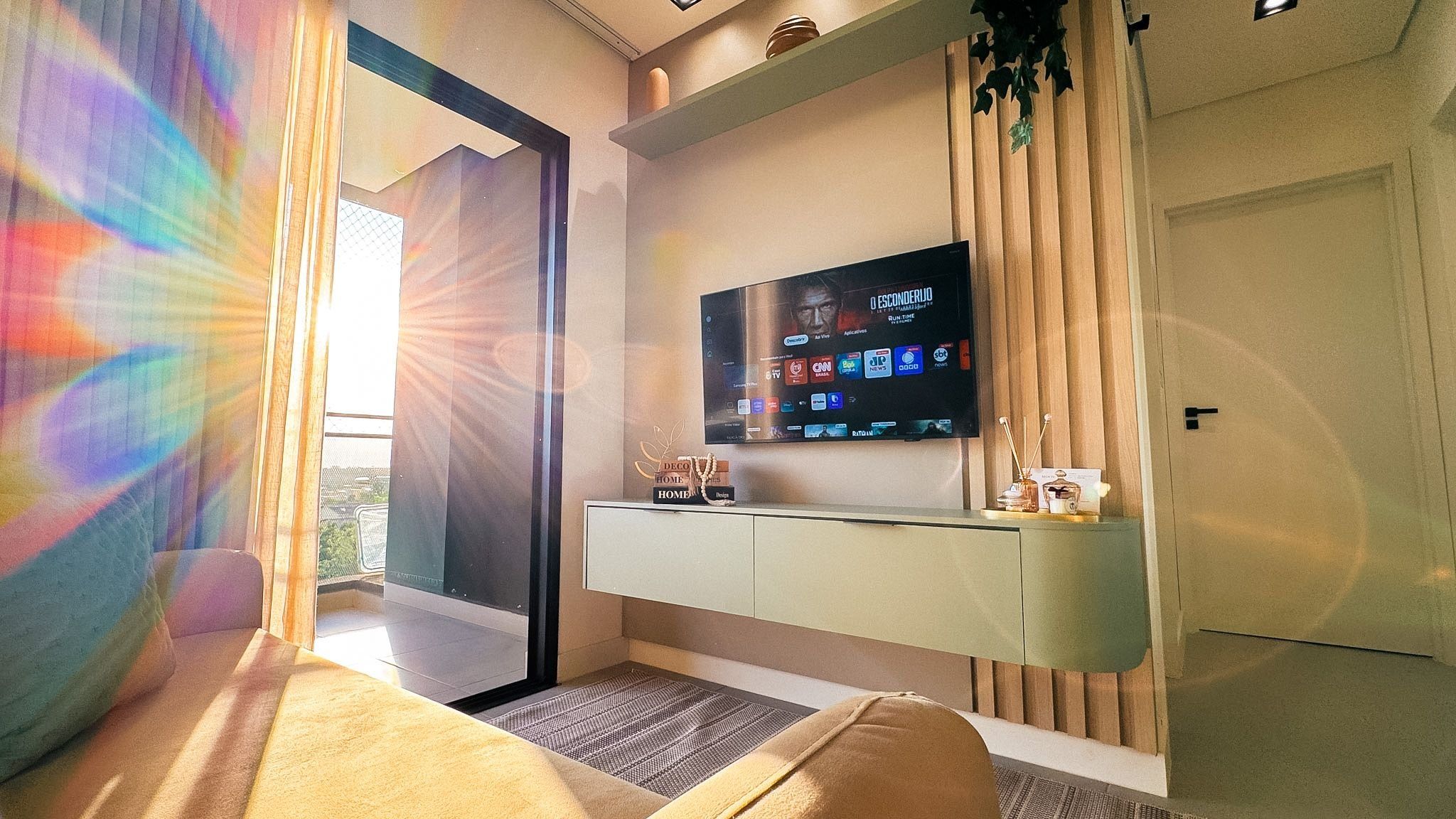 A modern hotel room with a large TV mounted on the wall, a seating area with a sofa, and a small table with decorative items.