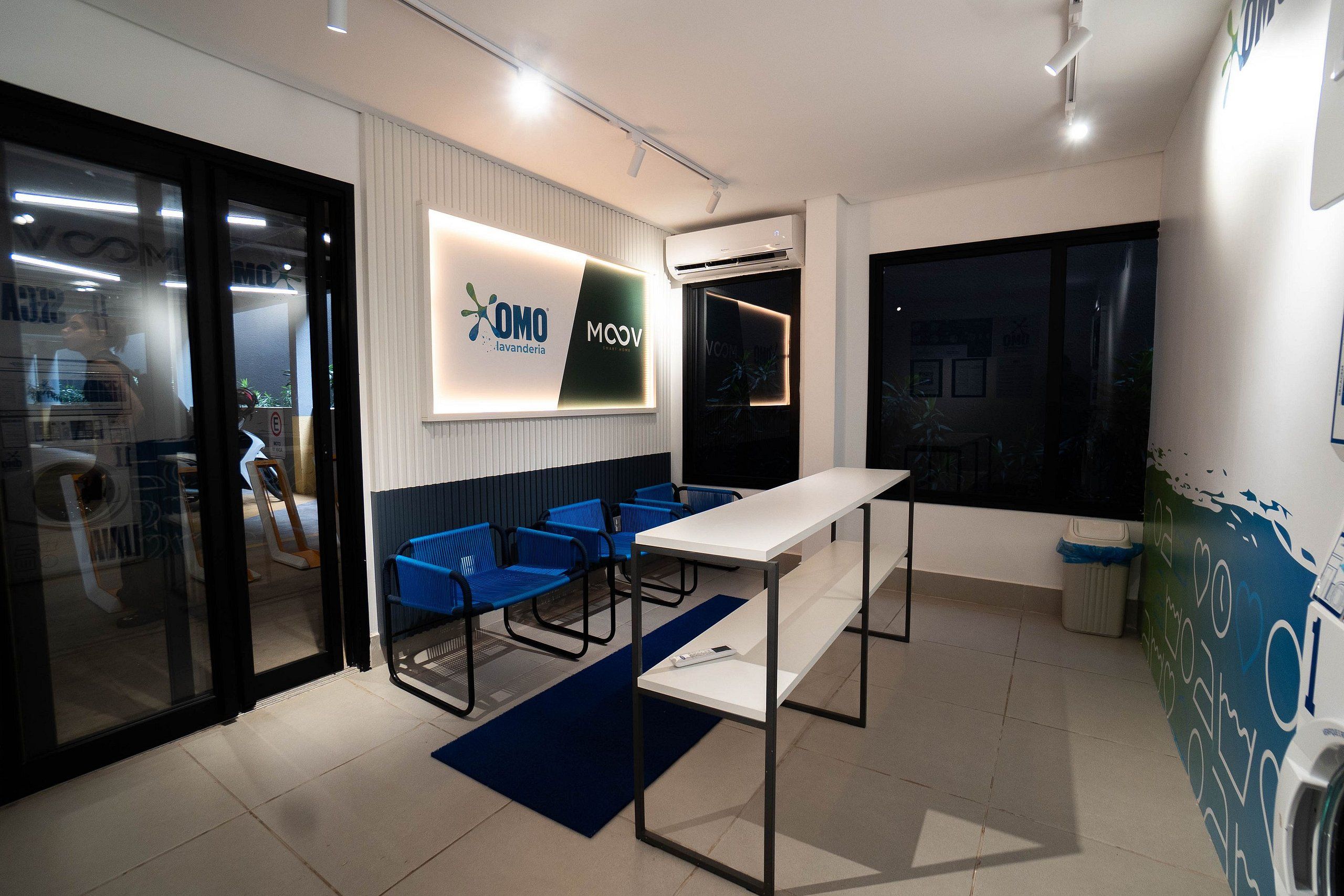 Un espacio de oficina moderno con zona de recepción, un mostrador blanco, sillas azules, un ventanal grande y una unidad de aire acondicionado montada en la pared.
