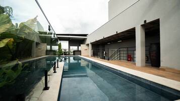 Una piscina con barrera de vidrio, una zona techada con techo y un edificio con un cartel 'MOV'.