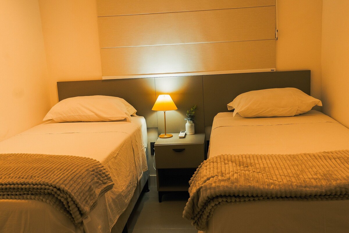 A hotel room with two single beds, a nightstand with a lamp, and a small table with a vase and a phone.